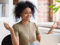 African young woman sitting at table opposite laptop in workplace meditating doing yoga exercise smiling feels good. Take a break, inner balance and harmony, stress relief no negative emotions concept (African young woman sitting at table opposite lap